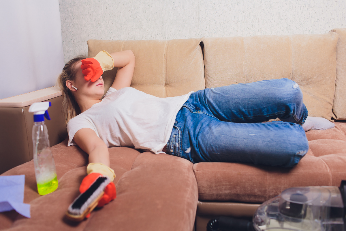 an exhausted woman wearing cleaning gloves laying on a brown sectional holding a bristle brush and upholstery cleaner